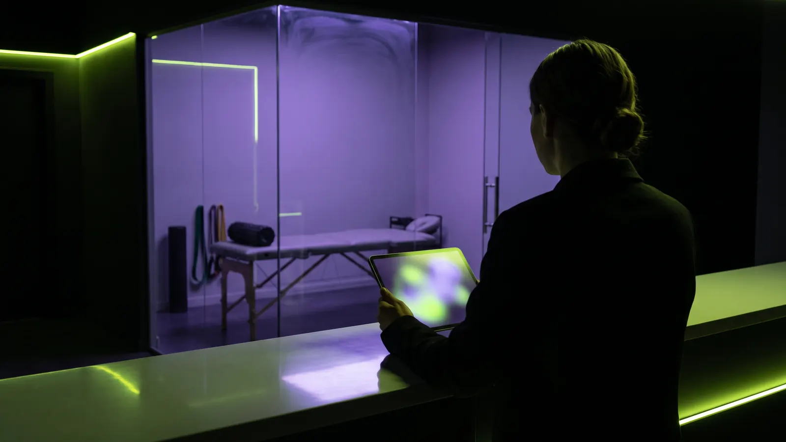 A regenerative clinic owner, seen from behind, holding a tablet at the front desk of an empty sports medicine clinic at night.
